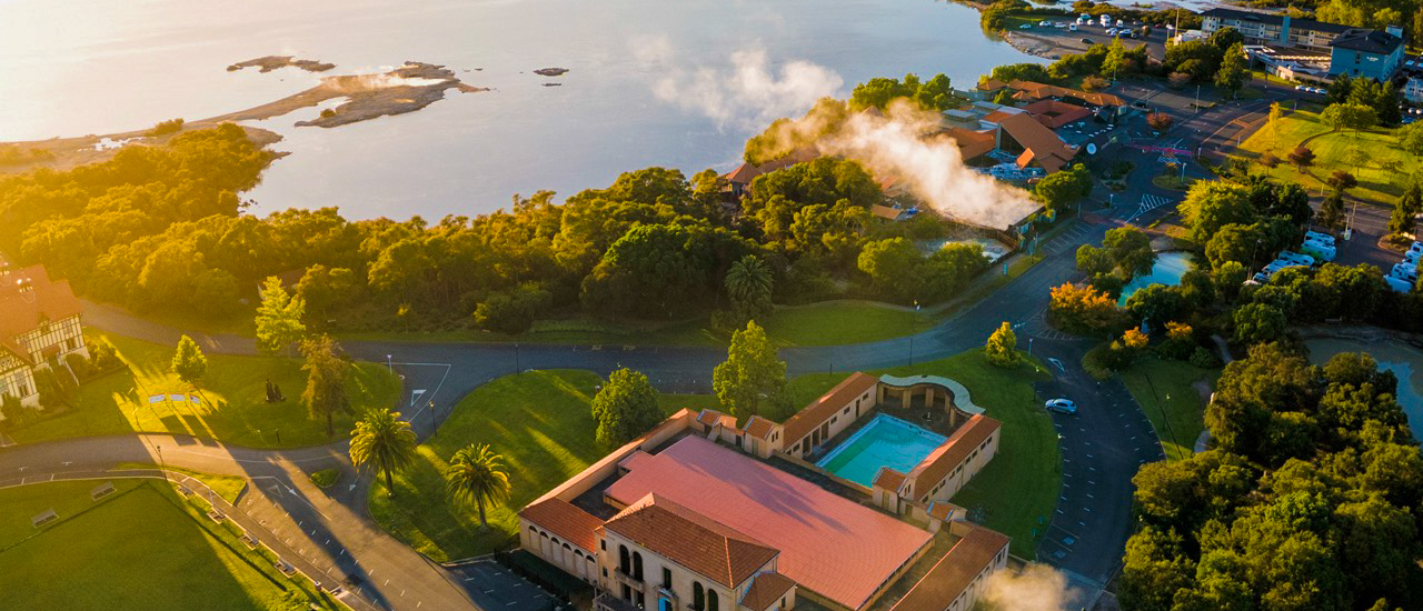 Golden light illuminates Rotorua, with geothermal steam rising from the lake near the historic bathhouse.