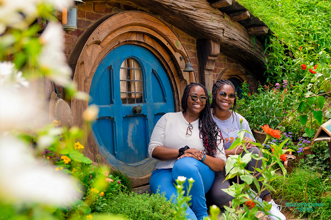 Two happy women smiling in front of a blue Hobbit hole at the magical Hobbiton film set.