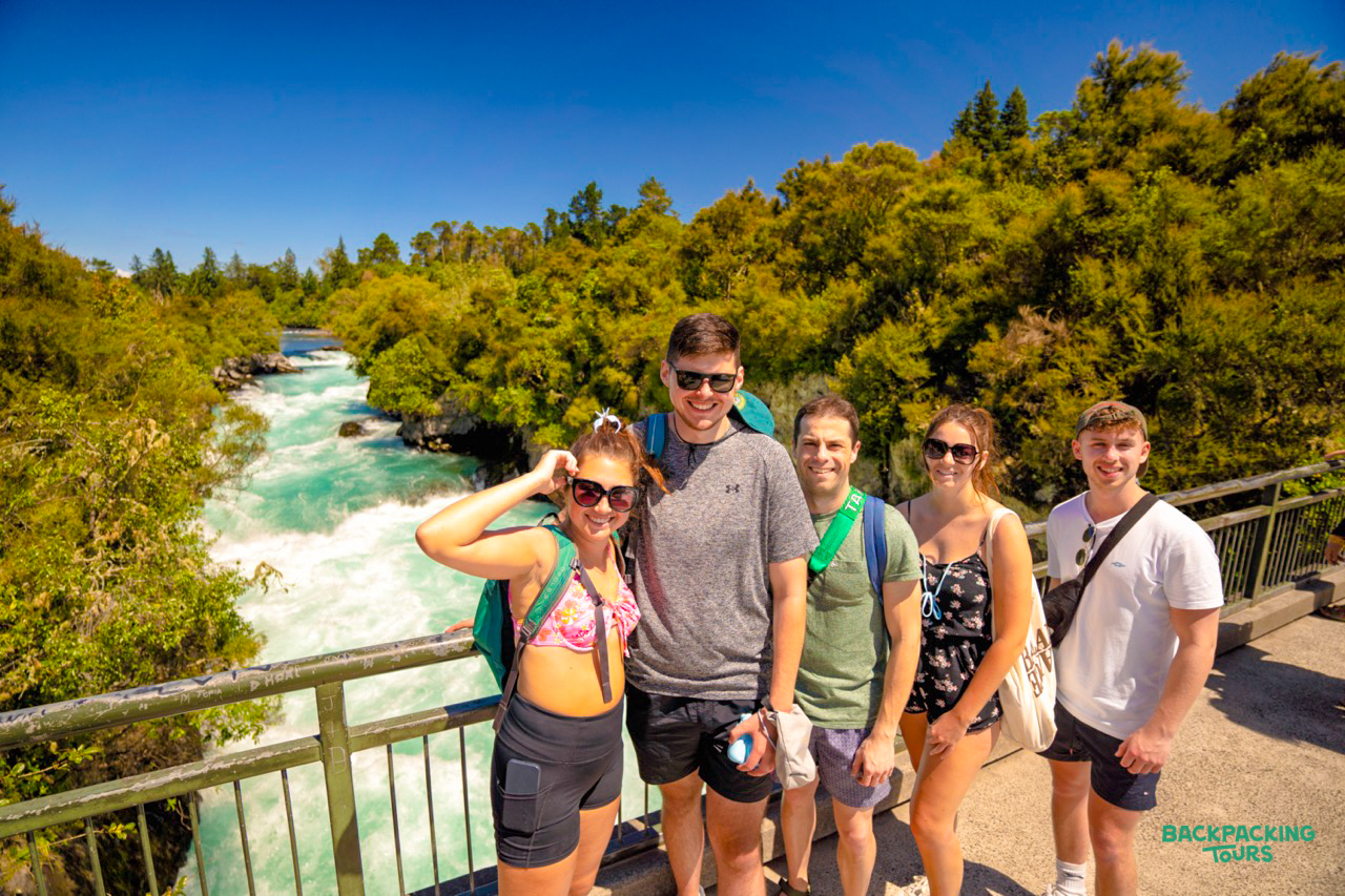 Five happy backpackers pose on a bridge overlooking the stunning turquoise Huka Falls in New Zealand.