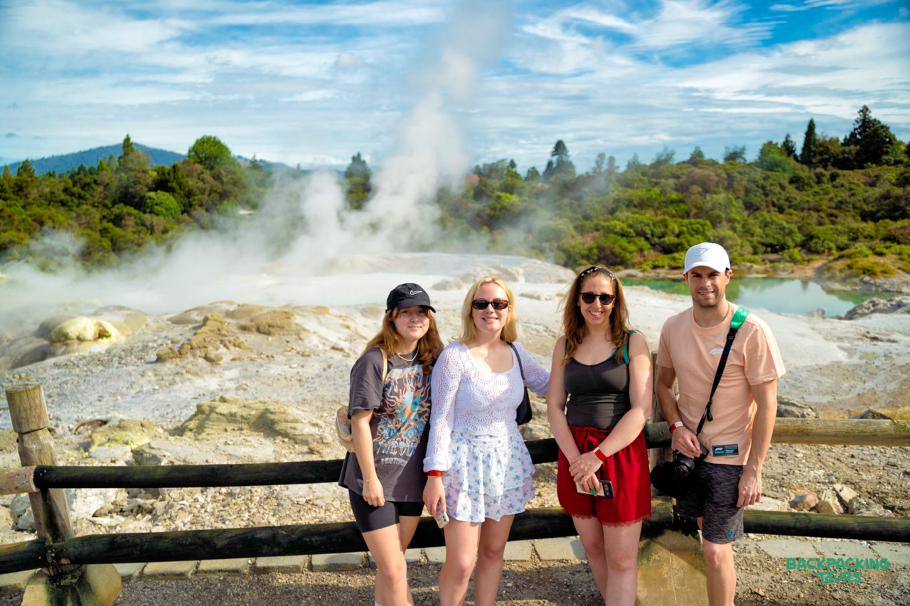 Four happy backpackers pose in front of an erupting geyser and steaming pools in Rotorua.