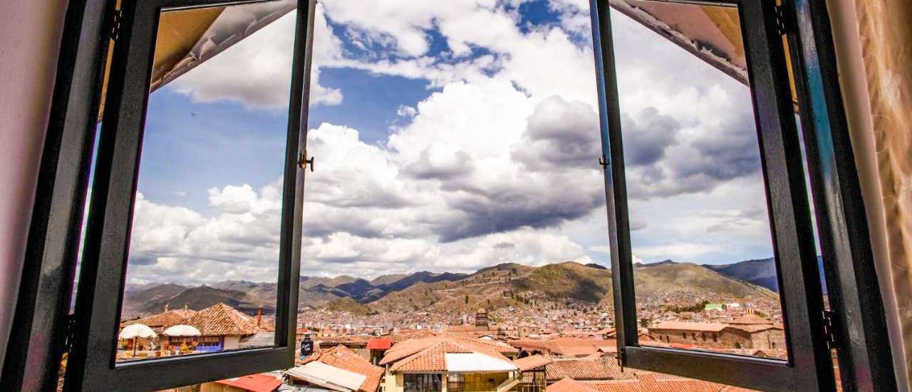 An open window frames a panoramic view of Cusco city with terracotta roofs and mountains under a cloudy sky.