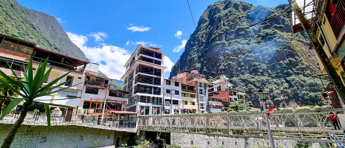 Bustling Aguas Calientes town with buildings and a bridge, surrounded by towering green mountains.