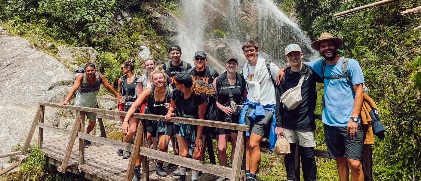 A happy group of hikers on a wooden bridge in front of a waterfall on the Inca Trail.