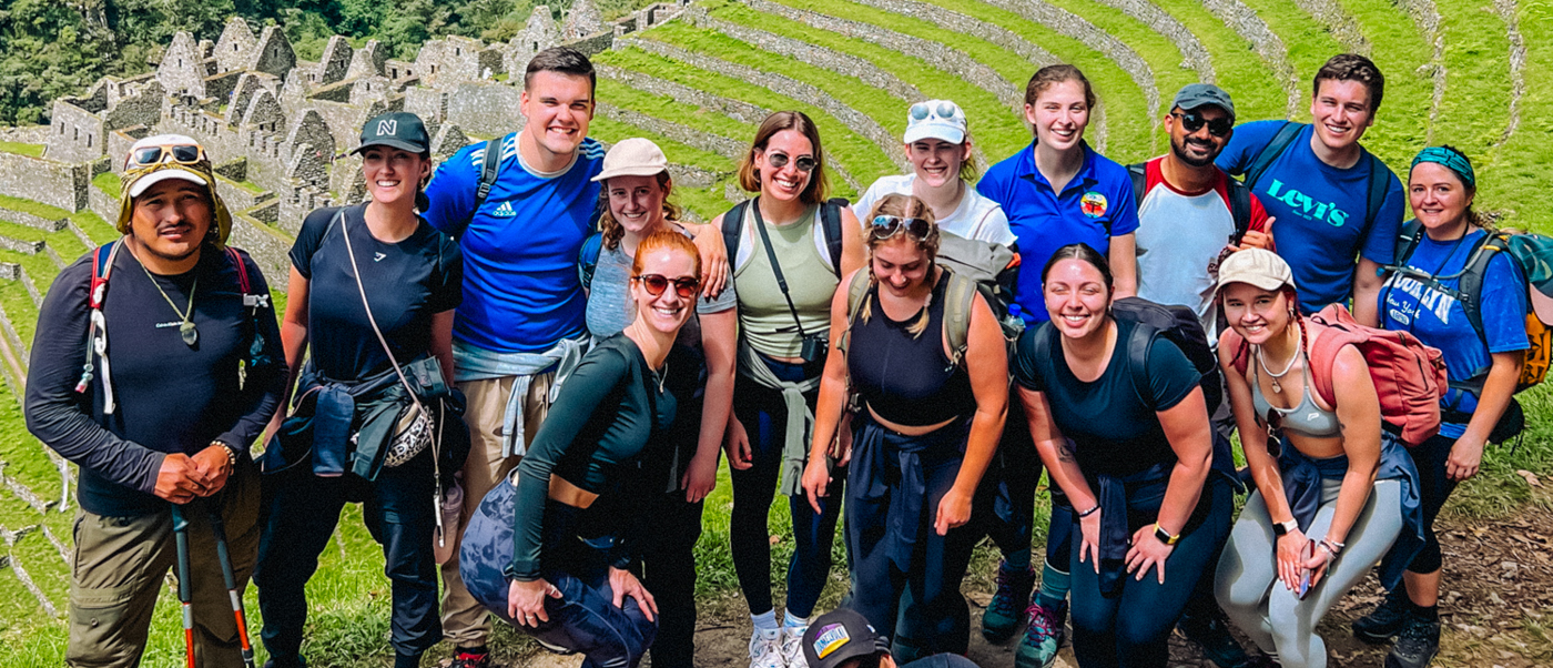 Smiling hikers on the Inca Trail in Peru, with ancient stone terraced ruins in the background.