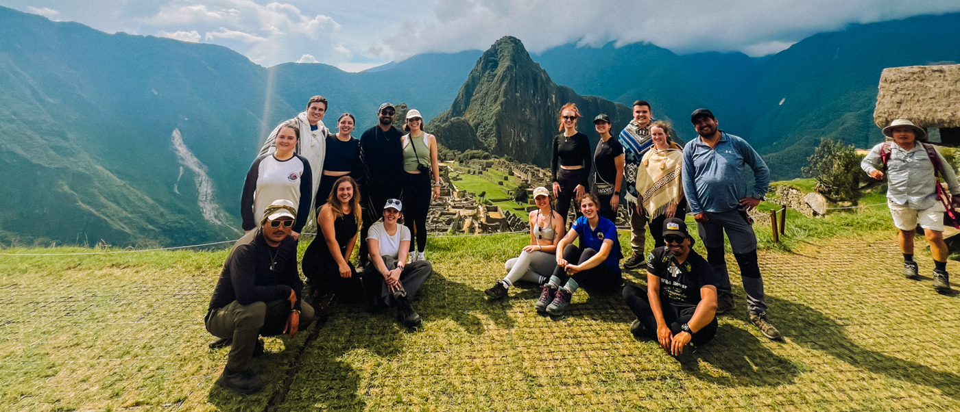 Smiling backpackers pose on a grassy hill overlooking the ancient city of Machu Picchu.