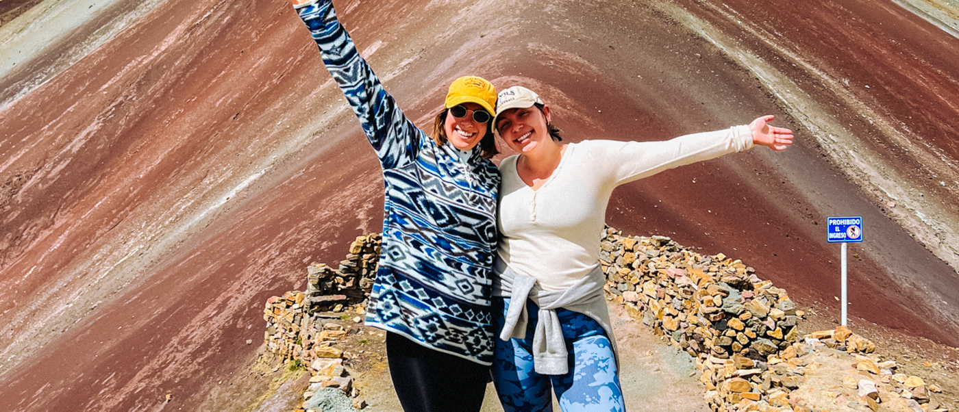 Two happy women pose with arms outstretched on a rocky path at Rainbow Mountain, Peru.