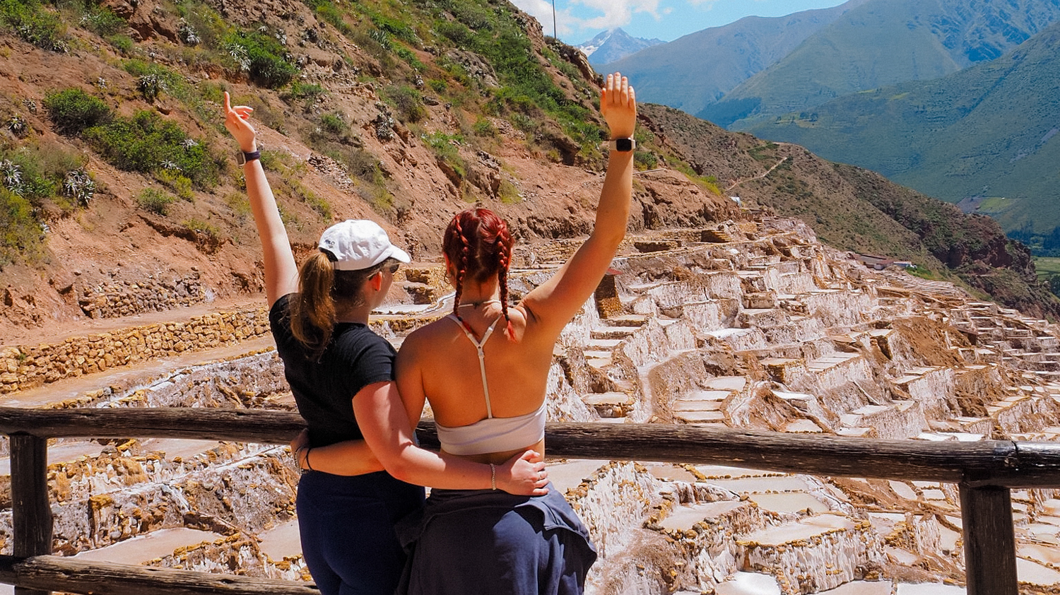 Two joyful women with raised arms embrace, overlooking the terraced Maras Salt Flats in Peru.
