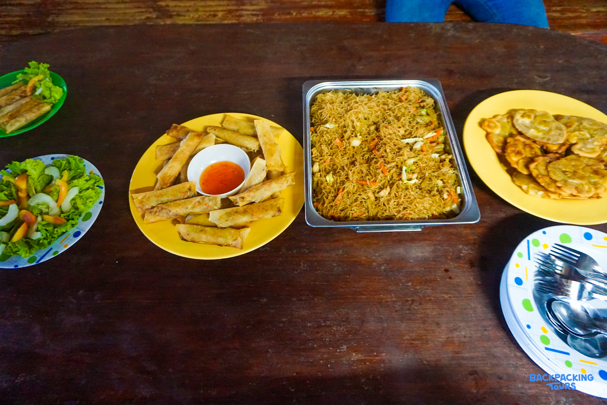 A delicious homemade Filipino lunch with spring rolls, noodles, and fresh salad on a wooden table.