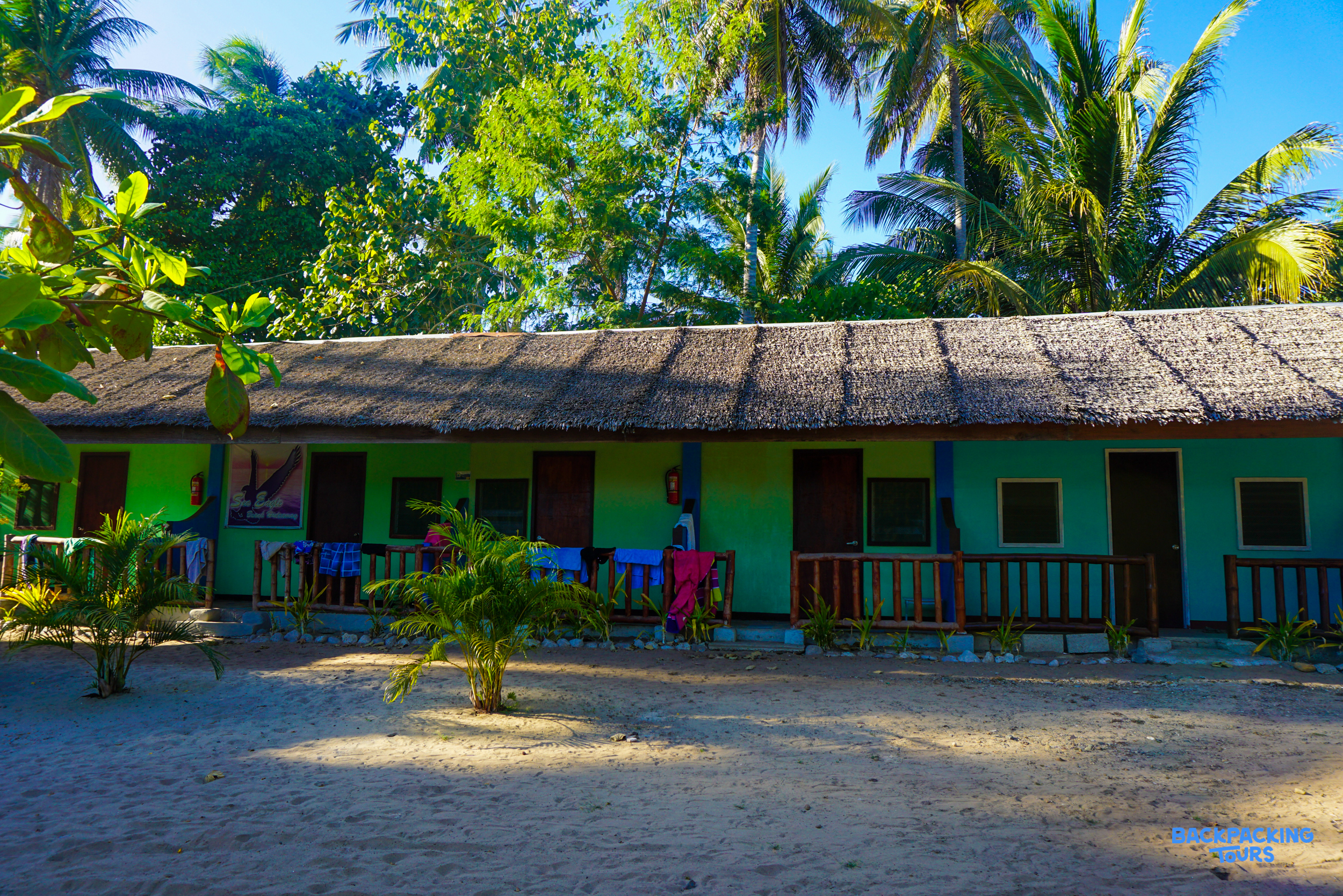 Colorful bungalows with thatched roofs on a sandy beach, surrounded by lush palm trees in the Philippines.