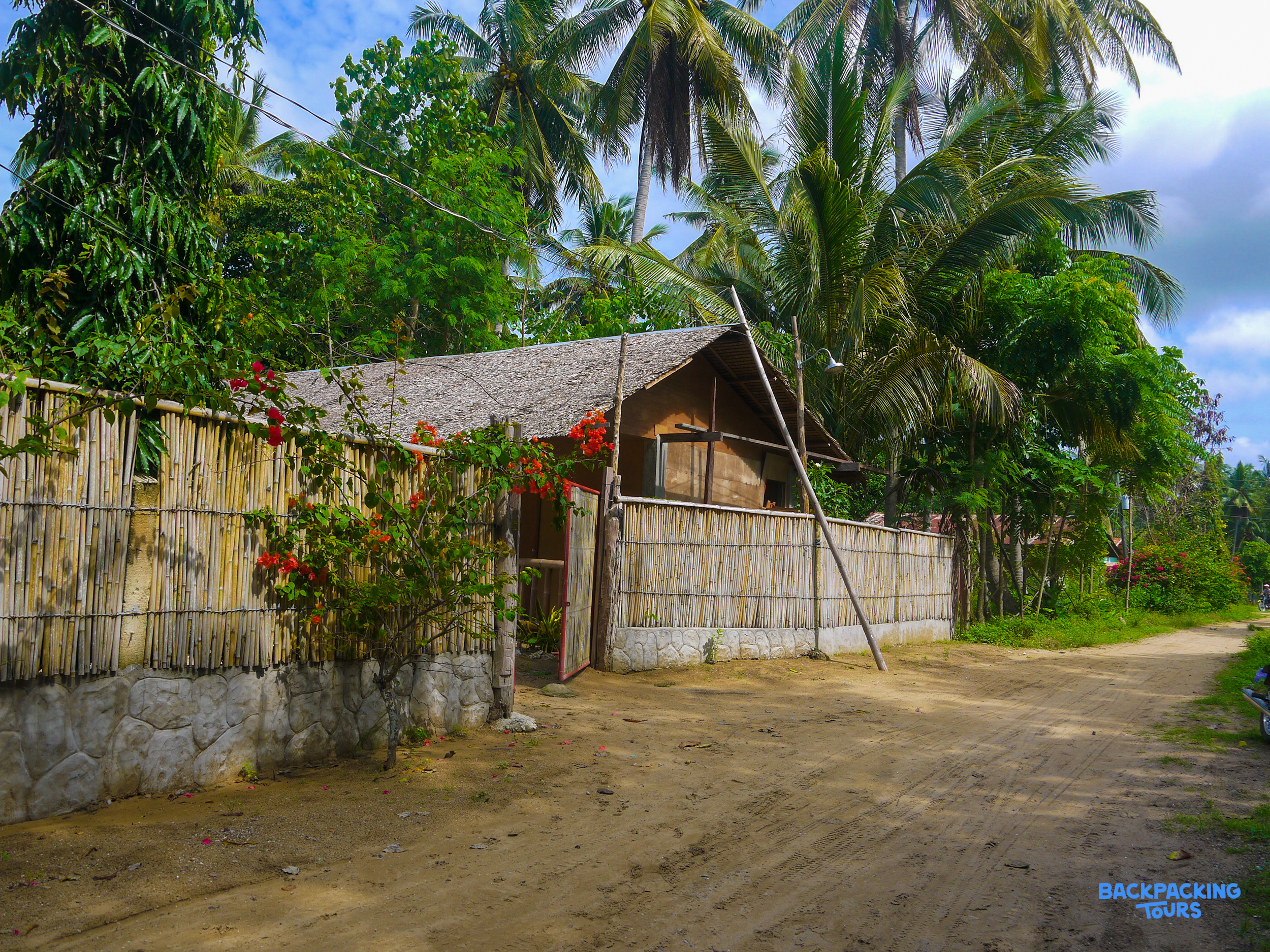 A simple tropical home with a thatched roof and bamboo fence, set among lush palm trees.
