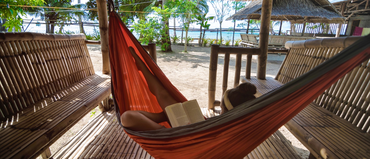A person relaxes in an orange hammock, reading a book with a tropical beach in the background.
