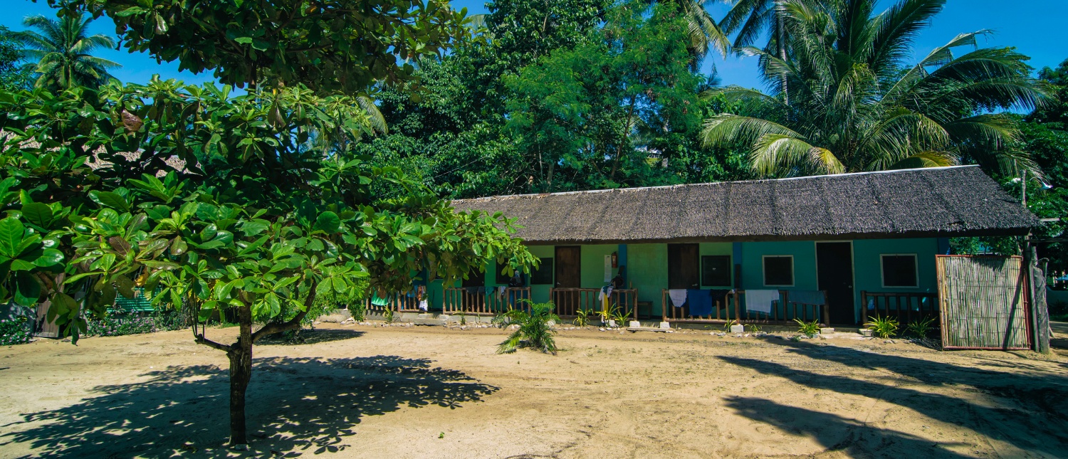Exterior of a tropical building with a thatched roof on sandy ground, surrounded by lush green trees.