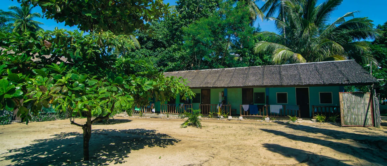 Exterior of a tropical building with a thatched roof on sandy ground, surrounded by lush green trees.