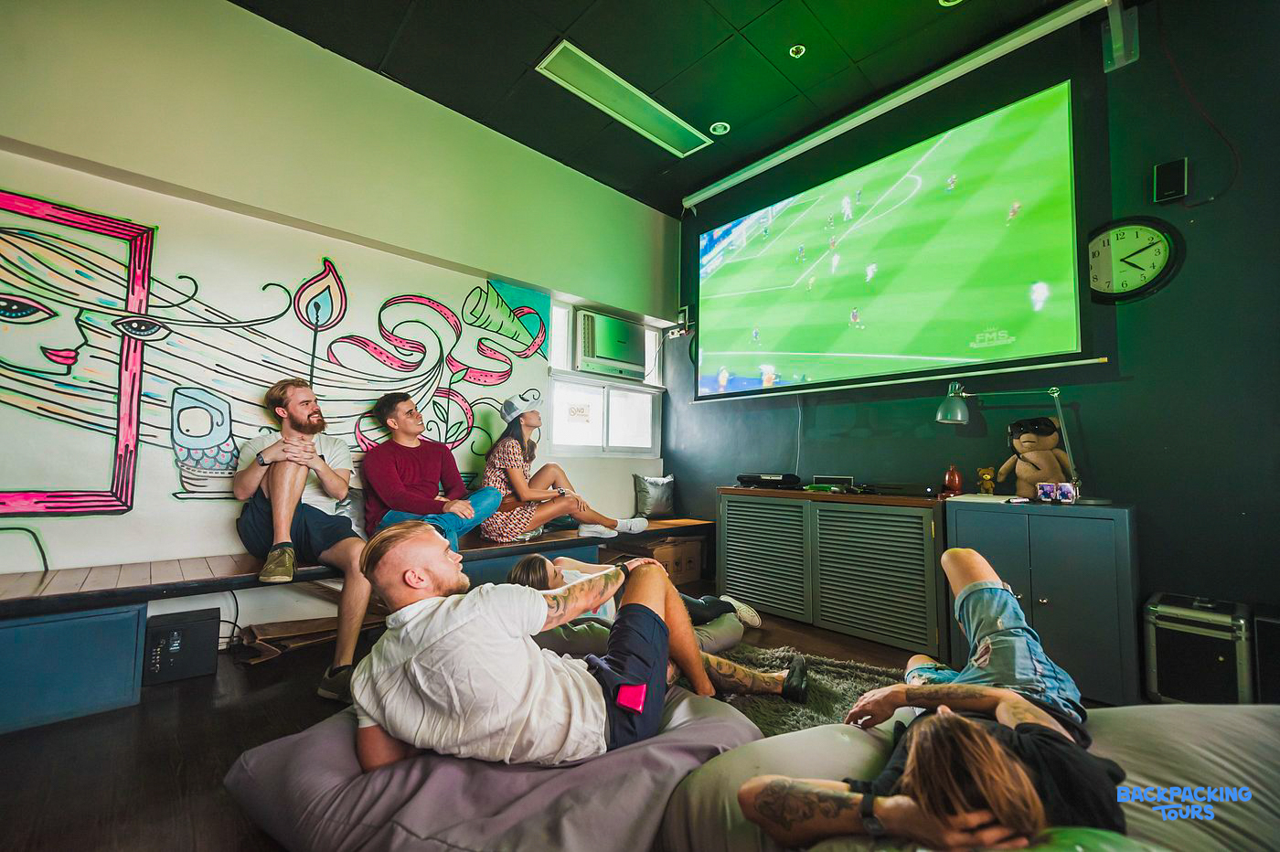 Backpackers relax on beanbags and a bench, watching a soccer game in a vibrant Manila hostel.