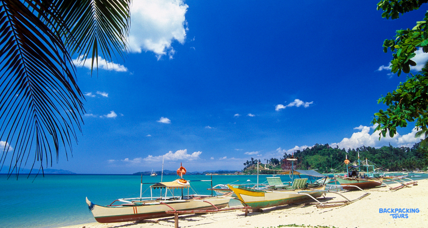 Outrigger boats line a beautiful white sand beach with turquoise water and lush green hills.