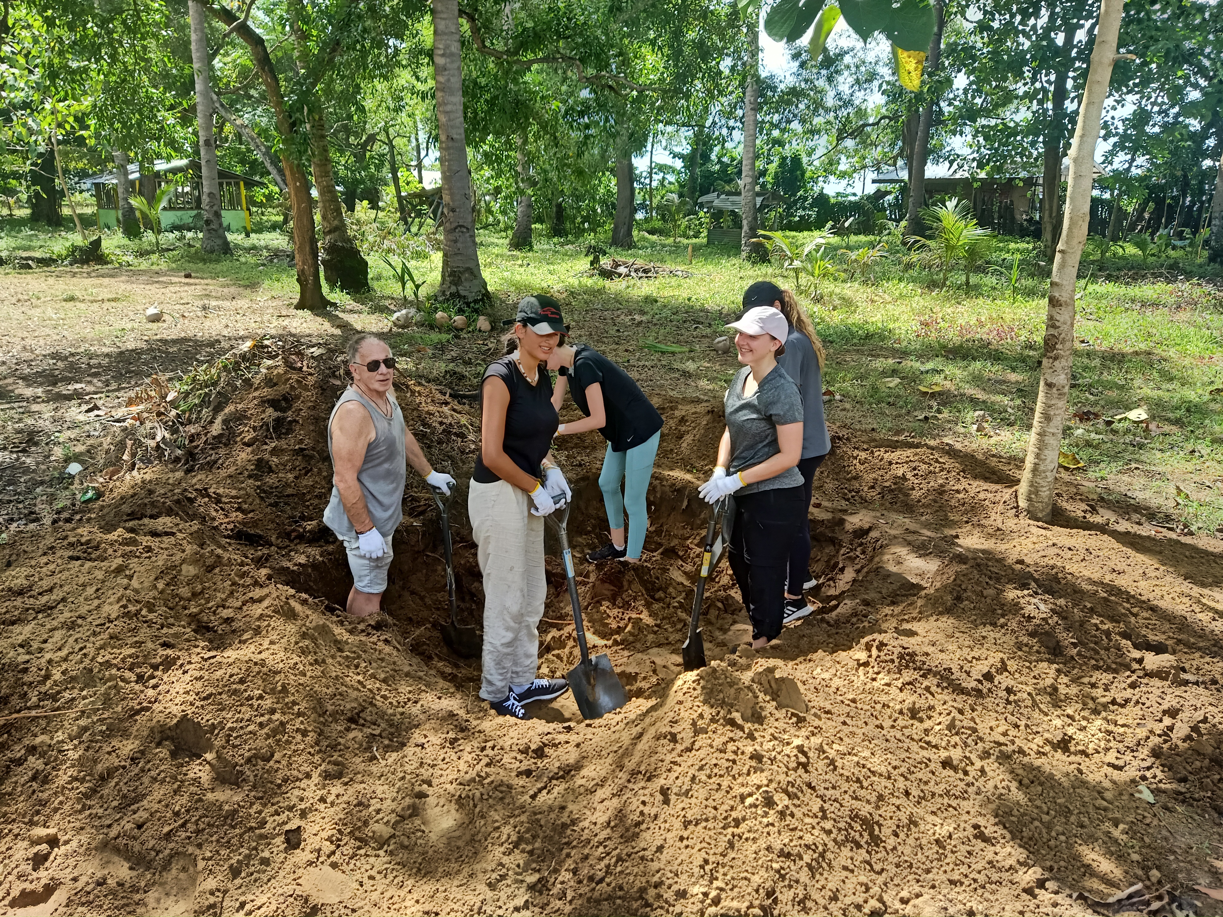 Five volunteers with shovels digging a large pit in the ground in a tropical Philippine village.