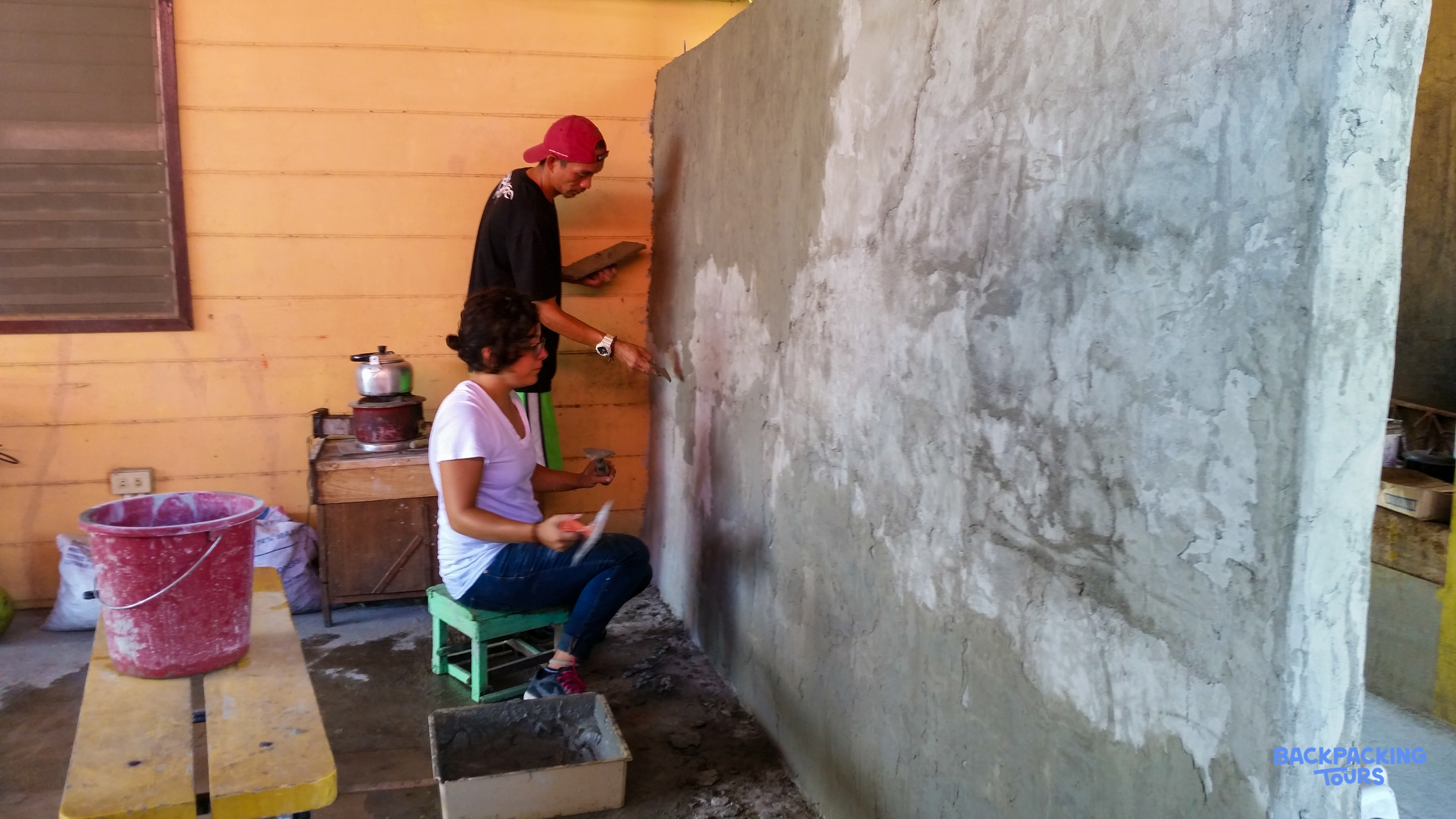 Two people carefully apply plaster to a large concrete wall during a renovation activity.