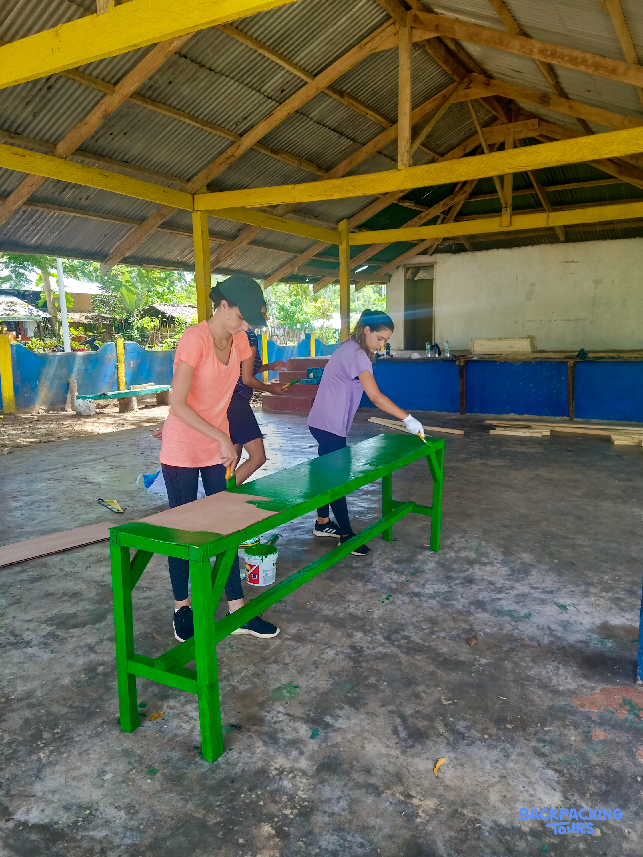 Two volunteers paint a long green wooden table under a yellow-beamed open-air shelter.