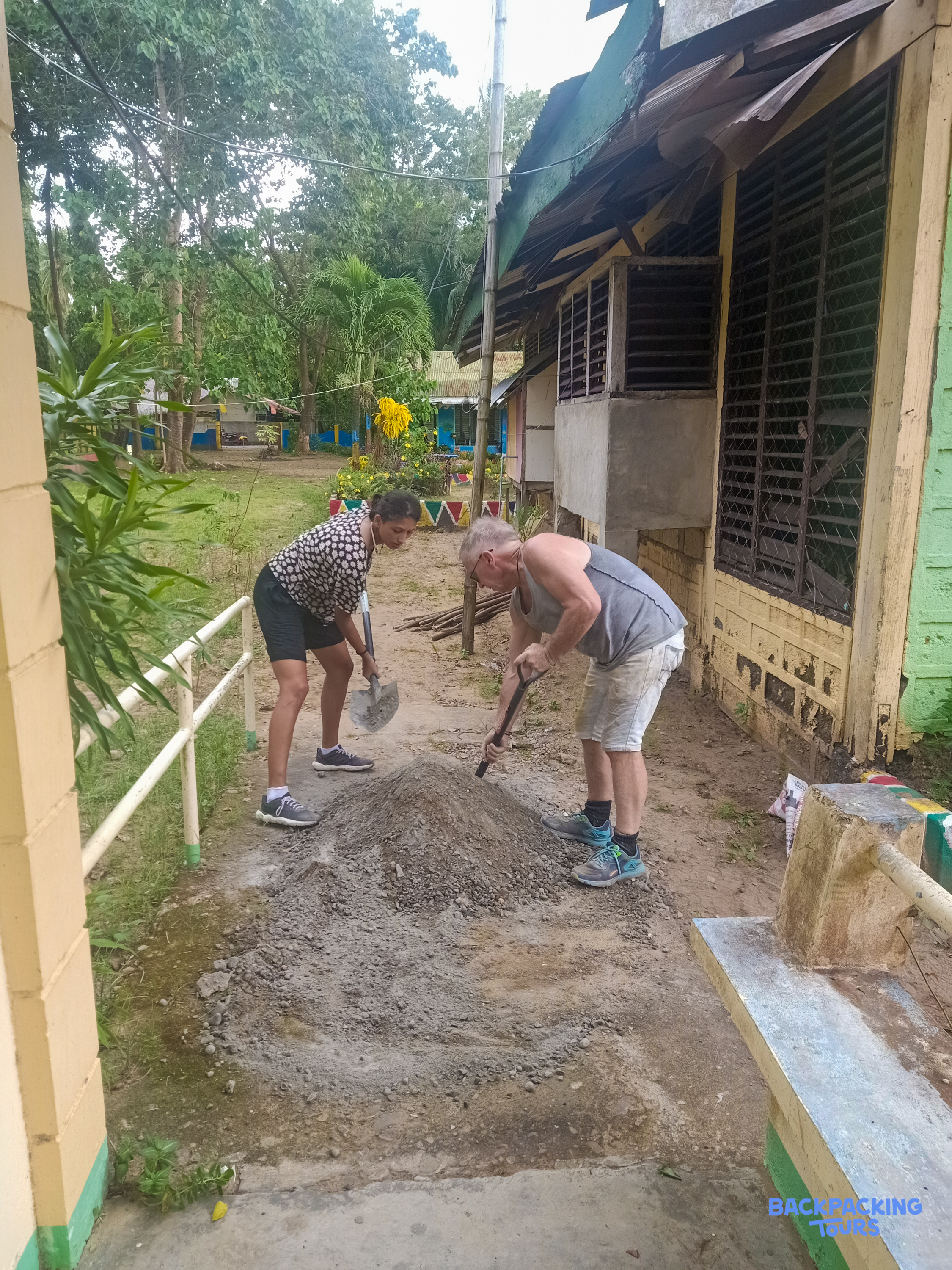 Two volunteers diligently mixing cement for a renovation project in a tropical outdoor setting.