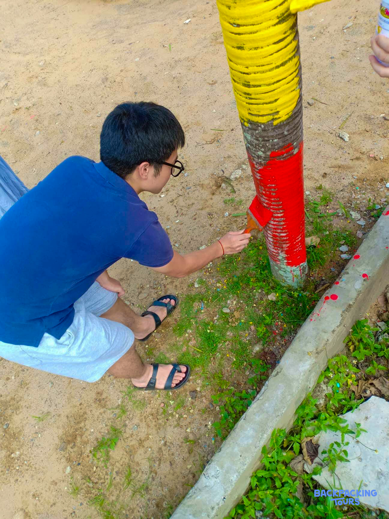 A person crouches, painting the base of a tree trunk red and yellow on sandy ground.