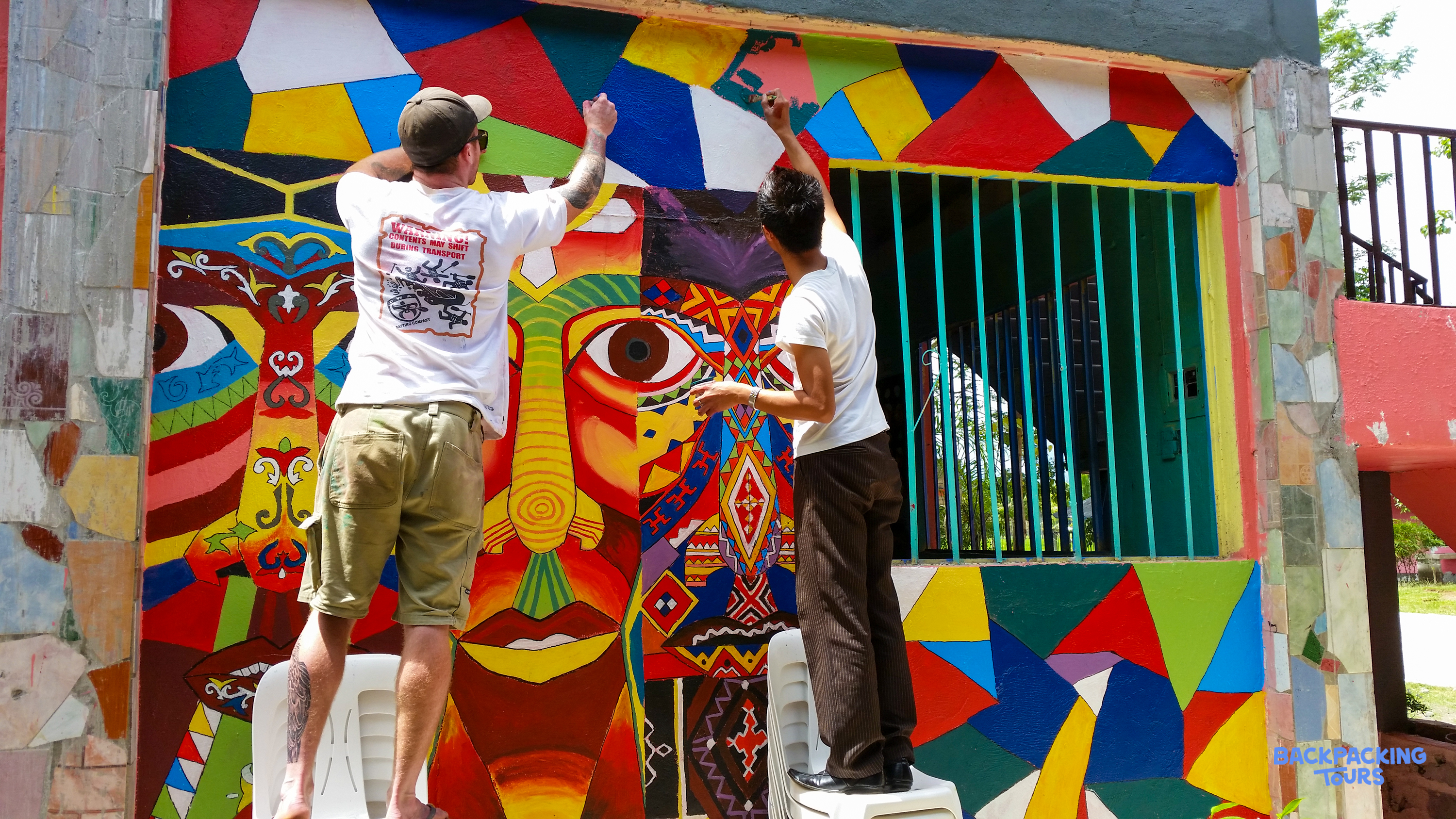 Two people creatively paint a bright, abstract mural featuring a large face on an outdoor wall.
