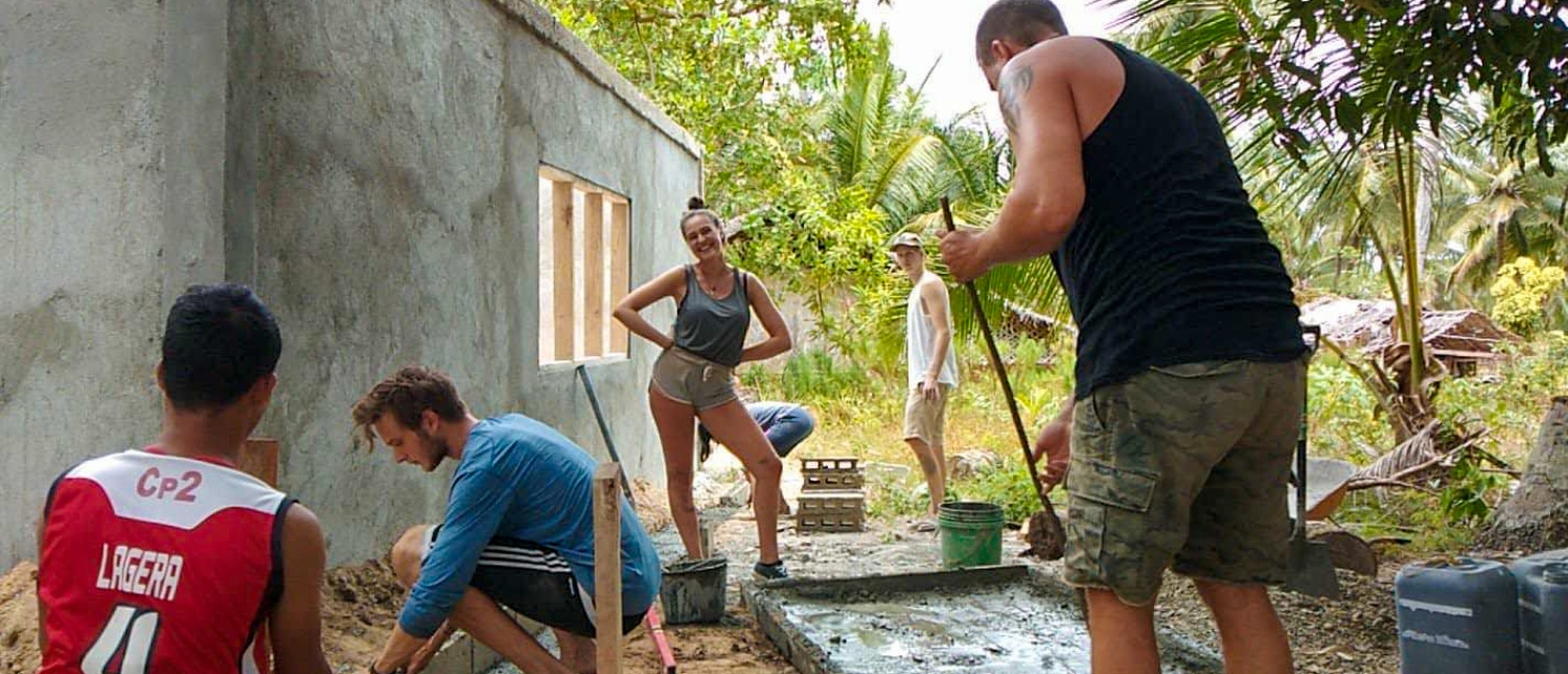 Smiling volunteers construct a building, mixing cement and laying blocks in a sunny, tropical setting.