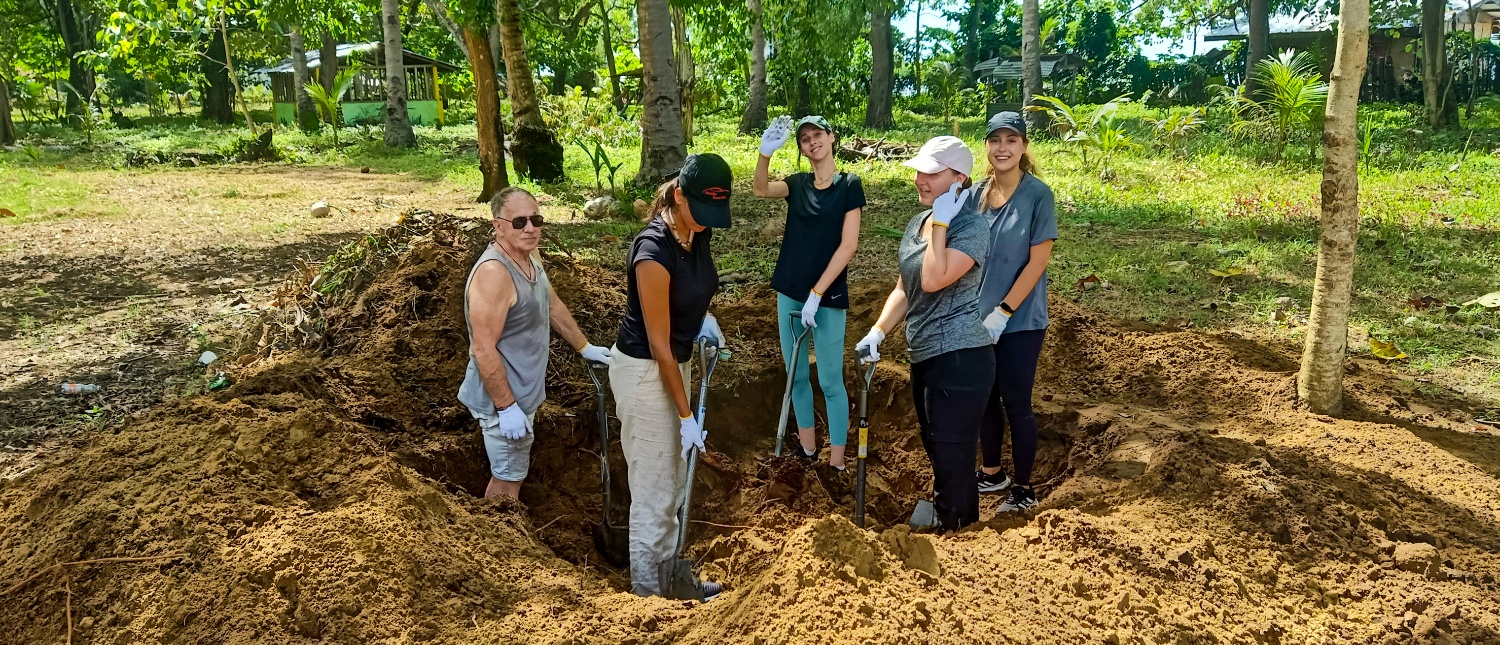 Five volunteers dig a large hole in the earth, surrounded by trees and green foliage.