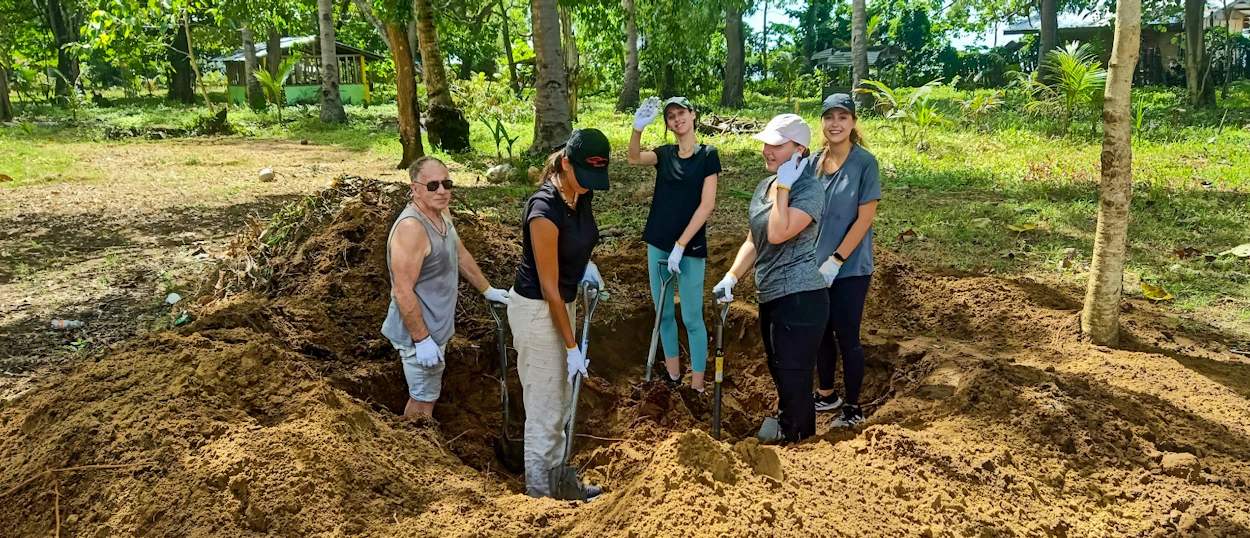 Five volunteers dig a large hole in the earth, surrounded by trees and green foliage.
