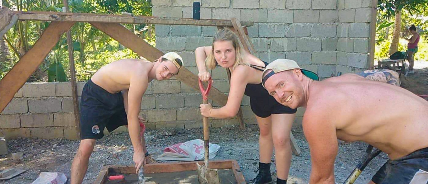 Three smiling volunteers mix cement for a building project in a tropical outdoor setting.