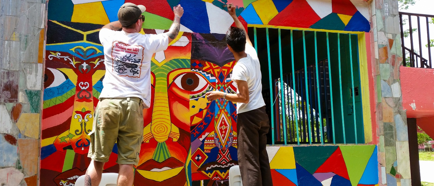 Two volunteers paint a vibrant, intricate mural with abstract faces and geometric patterns on a building.