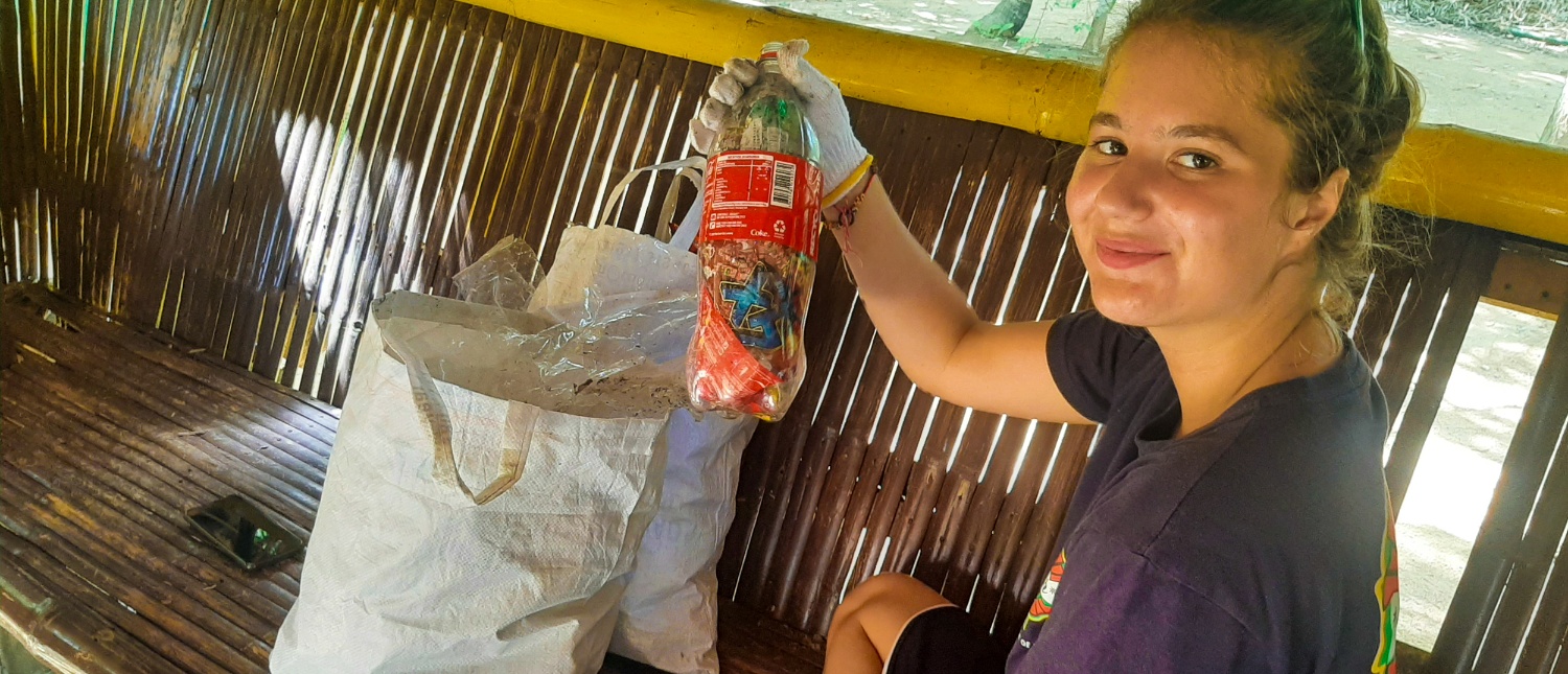 A smiling woman in gloves holds a bottle of collected beach trash, sitting on a bamboo bench.