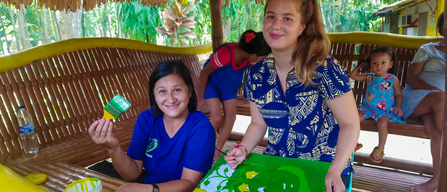 Two smiling women painting green designs on fabric in a bamboo hut in the Philippines.