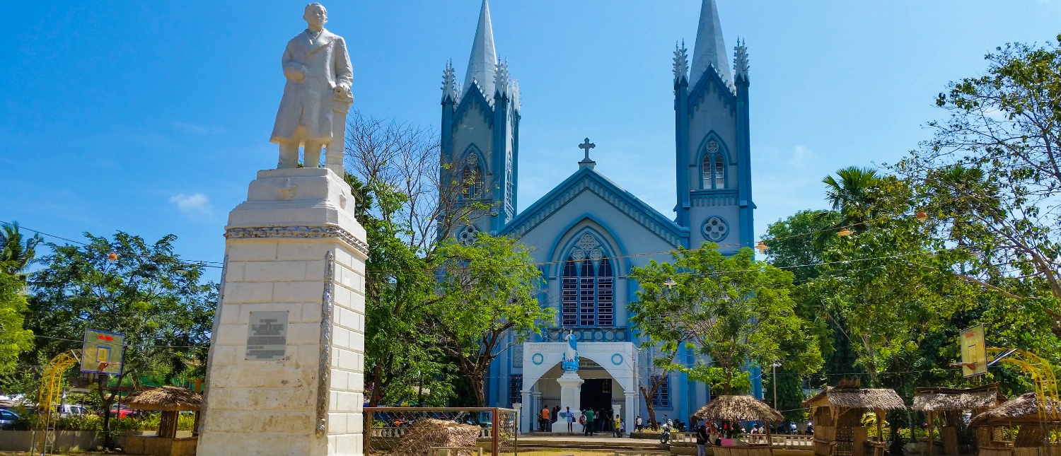 A grand light blue church with two spires behind a white statue in sunny Puerto Princesa.