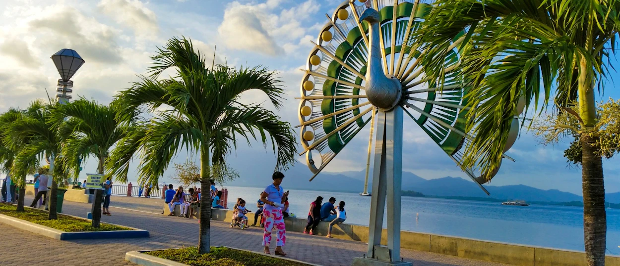 Palm trees line a sunny waterfront promenade in Puerto Princesa, with a peacock sculpture and strolling people.