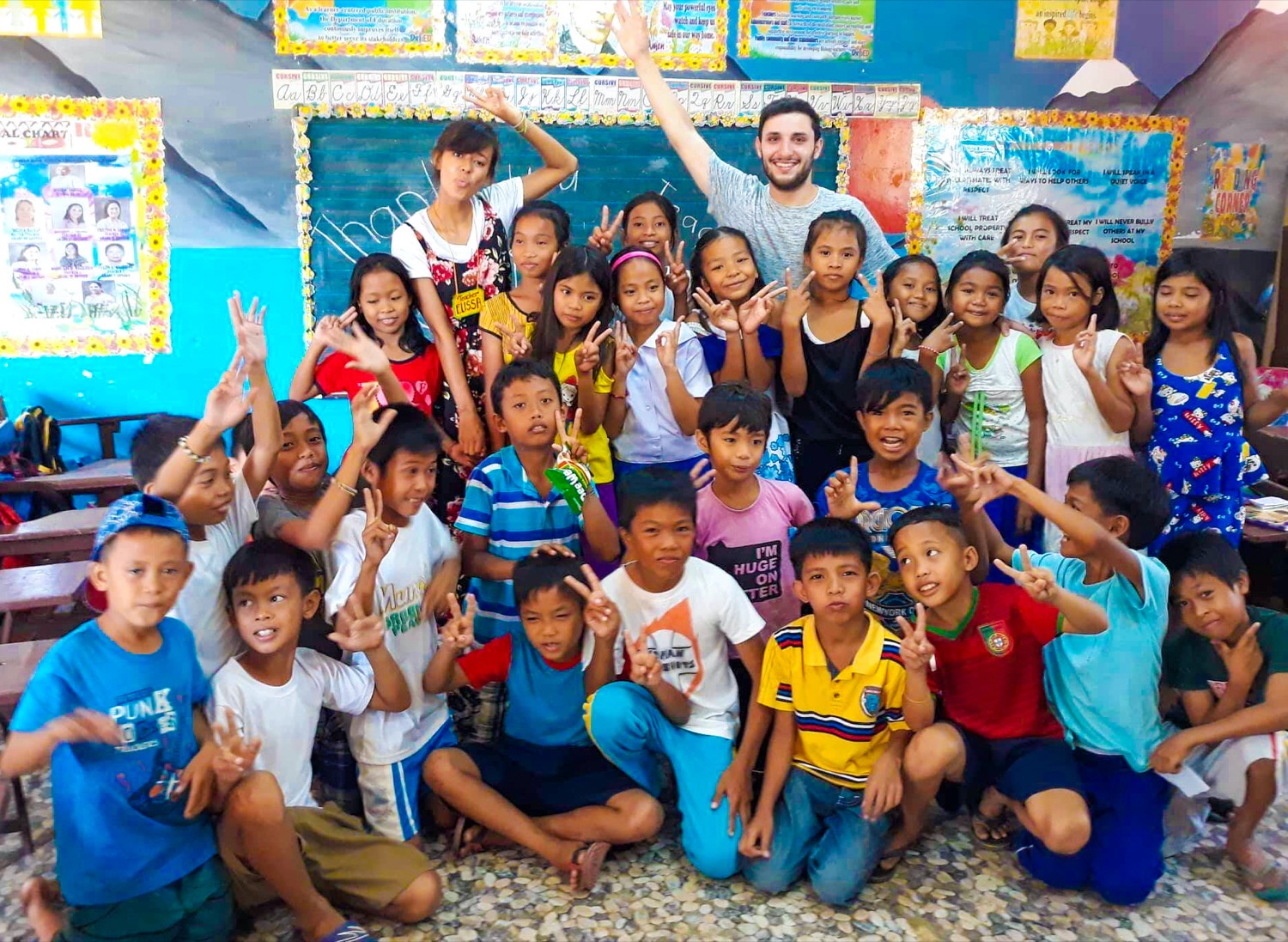 A joyful group of Filipino children and two teachers smiling and making peace signs in a classroom.