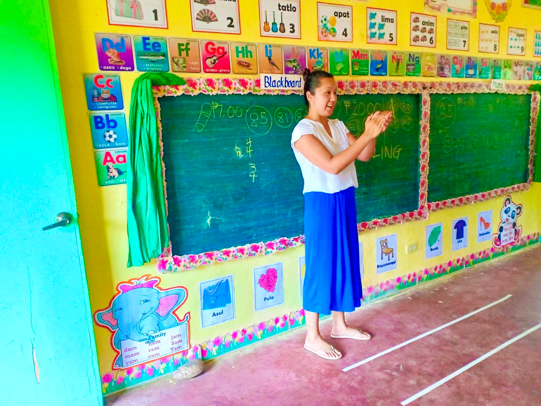 A smiling teacher stands barefoot in a brightly decorated classroom, ready to teach in Palawan.