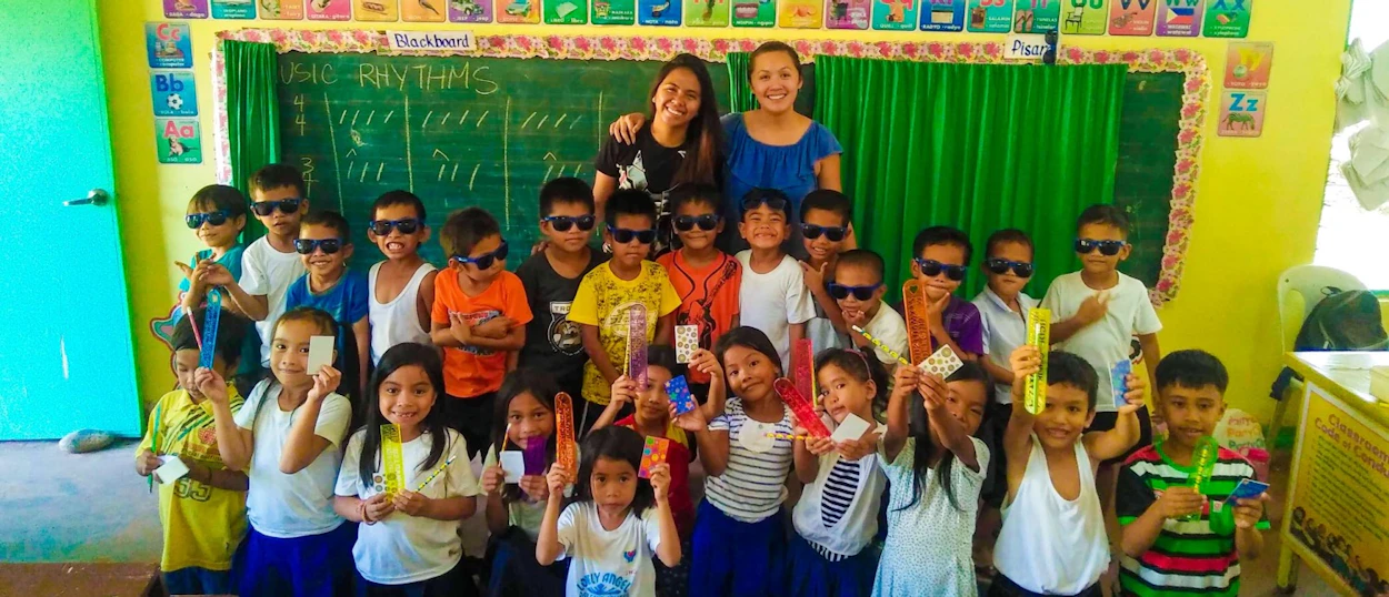 Smiling teachers and many children wearing sunglasses, holding colorful items, in a bright classroom.