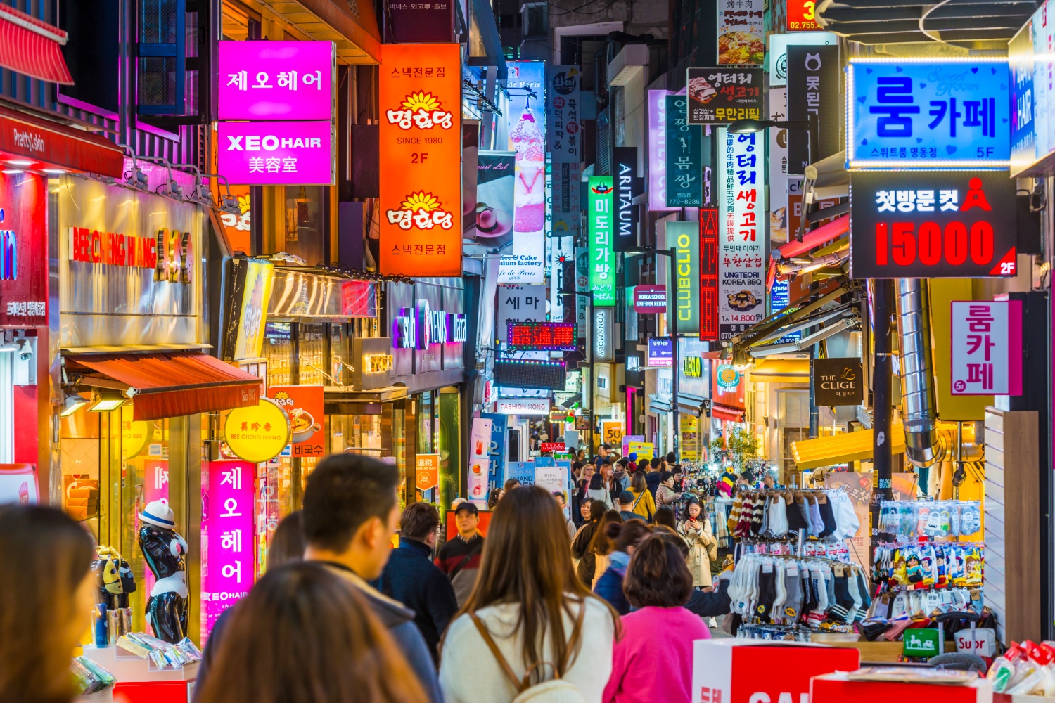 Crowds of people walk through a vibrant Myeongdong street at night, illuminated by colorful neon signs.