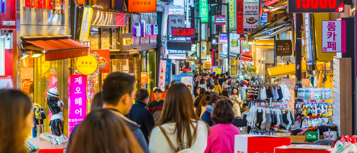 A vibrant, crowded shopping street in Seoul with many people and colorful neon signs.