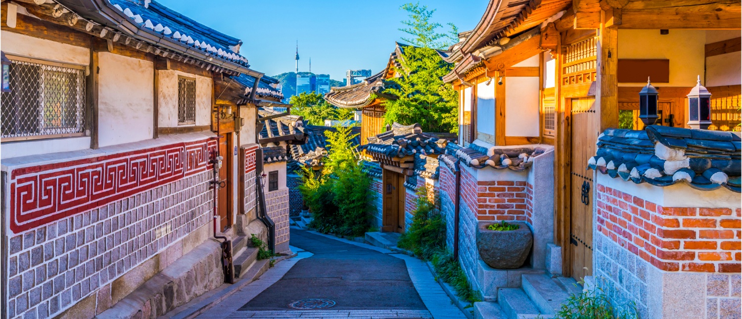 A charming traditional Korean Hanok village street, featuring tiled roofs, ornate walls, and green trees.