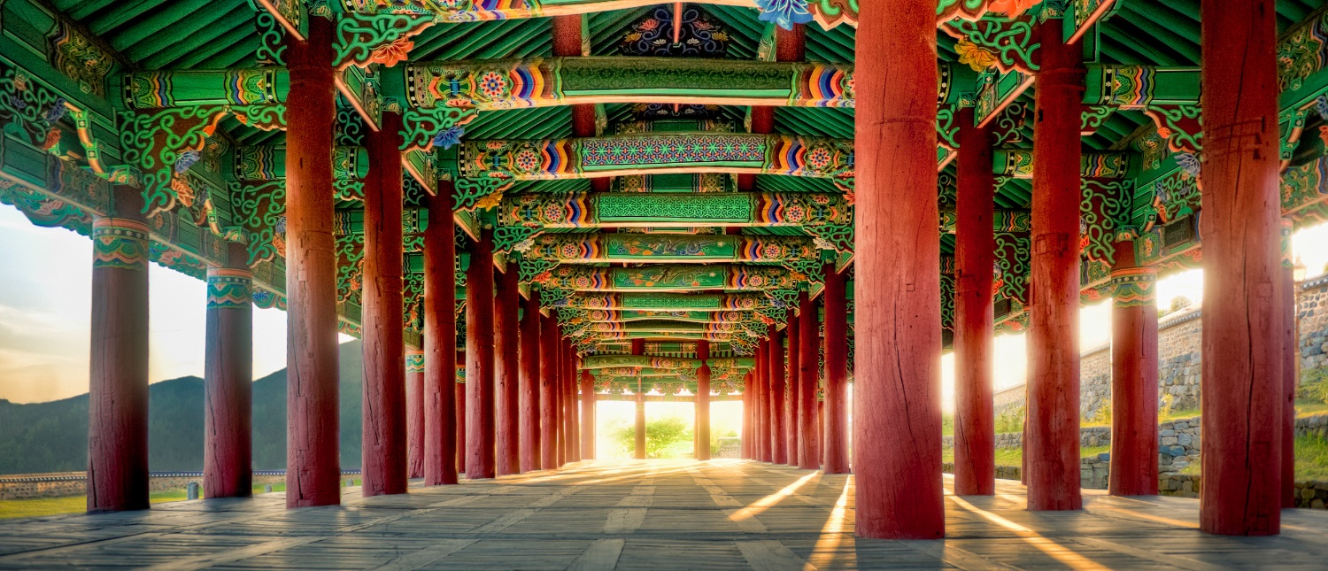 A long, vibrant corridor with red pillars and green decorated ceiling in Gyeongju, illuminated by sunlight.