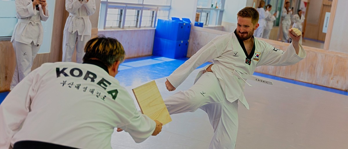 A taekwondo master holding a board while a student in a white dobok lands a flying kick.