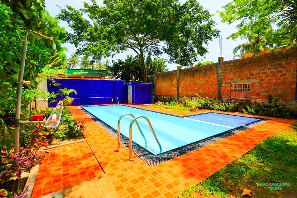A bright blue swimming pool with red tiled surround, lush green plants, and a rustic brick wall.