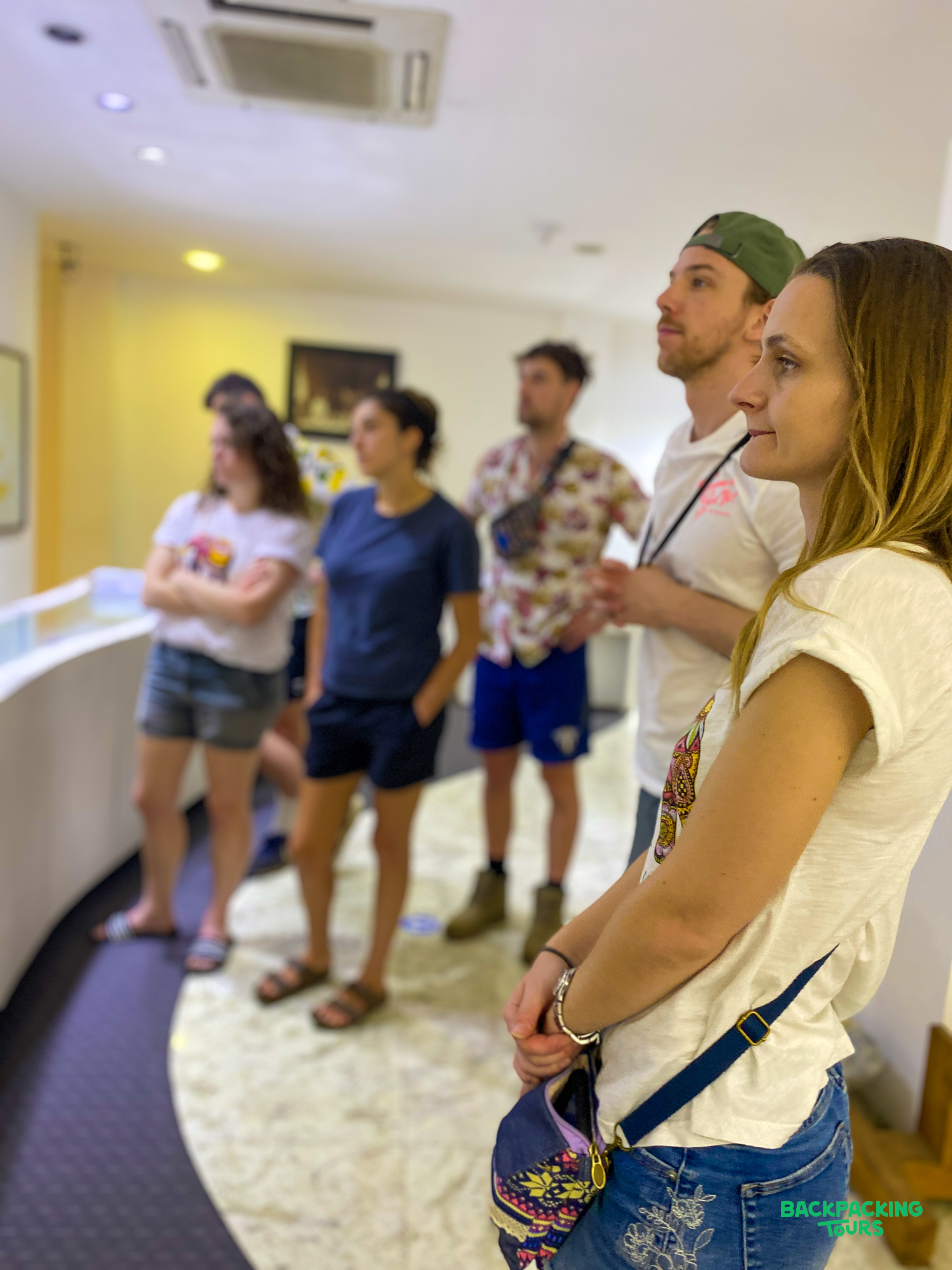 A group of young travelers attentively observe an unseen exhibit inside a modern building.