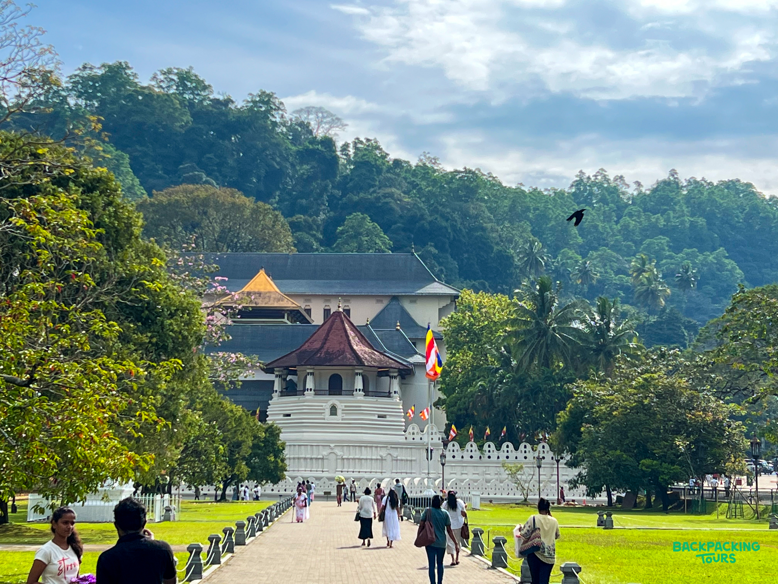 Visitors approach the stunning Temple of the Sacred Tooth Relic in Kandy, surrounded by lush tropical trees.