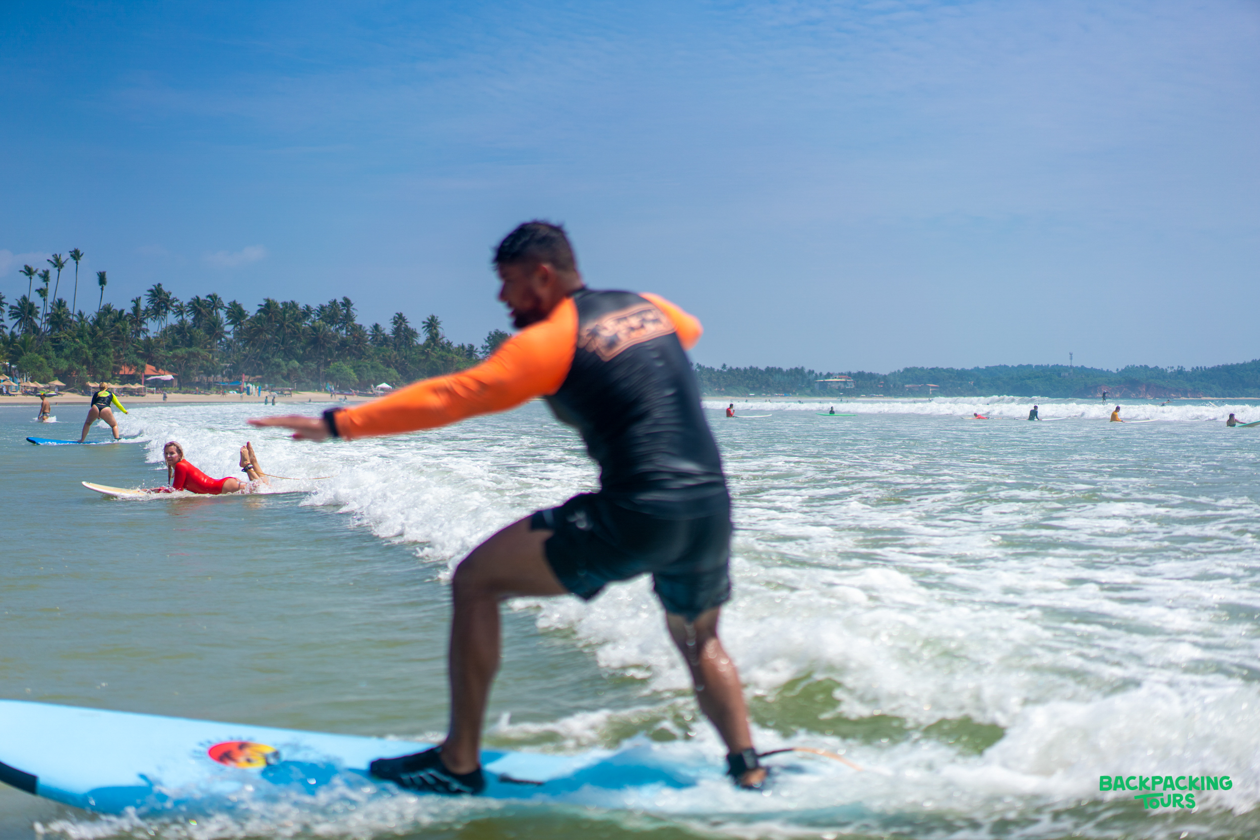 A man learns to surf on a blue board, balancing on a wave at sunny Weligama Beach.