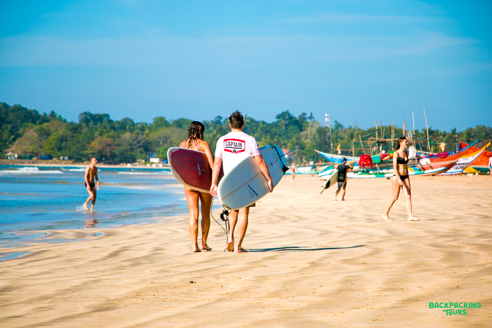 A couple walks along Mirissa beach with their surfboards under a sunny blue sky.