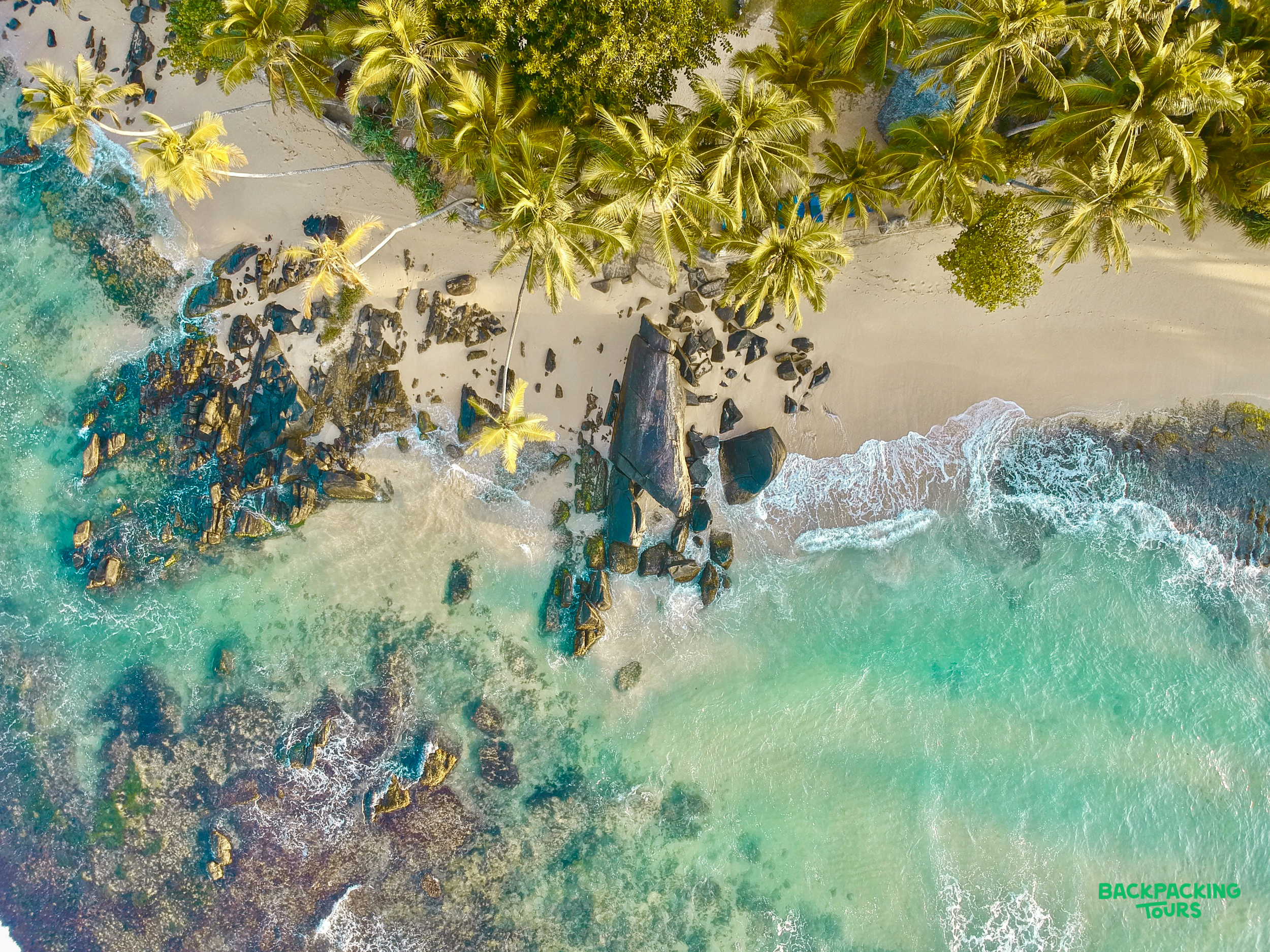 Aerial view of Weligama Beach, Sri Lanka, with clear turquoise waters, a rocky shore, and vibrant palm trees.