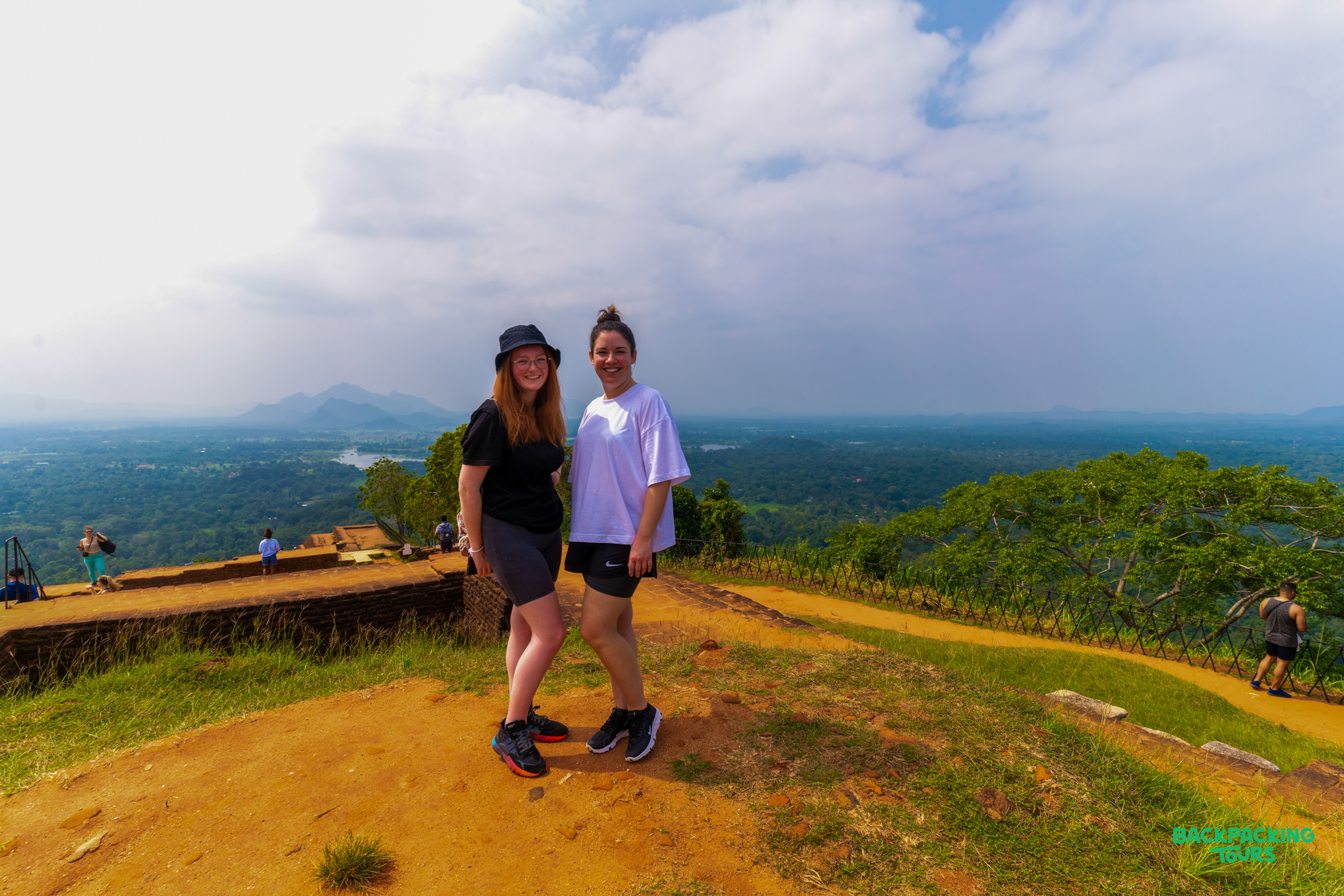 Two women pose on Sigiriya Rock, enjoying the expansive green landscape and cloudy sky.