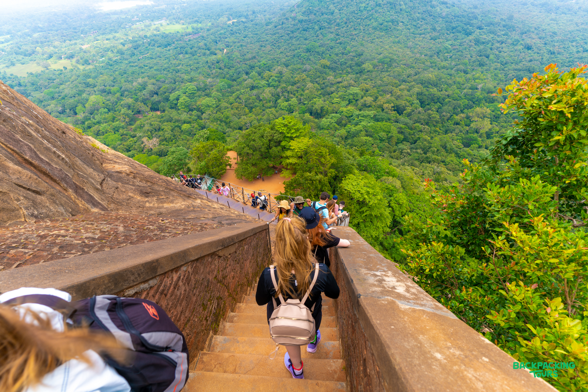 People descend the steep stone staircase at Sigiriya Lion Rock, with lush green forests below.